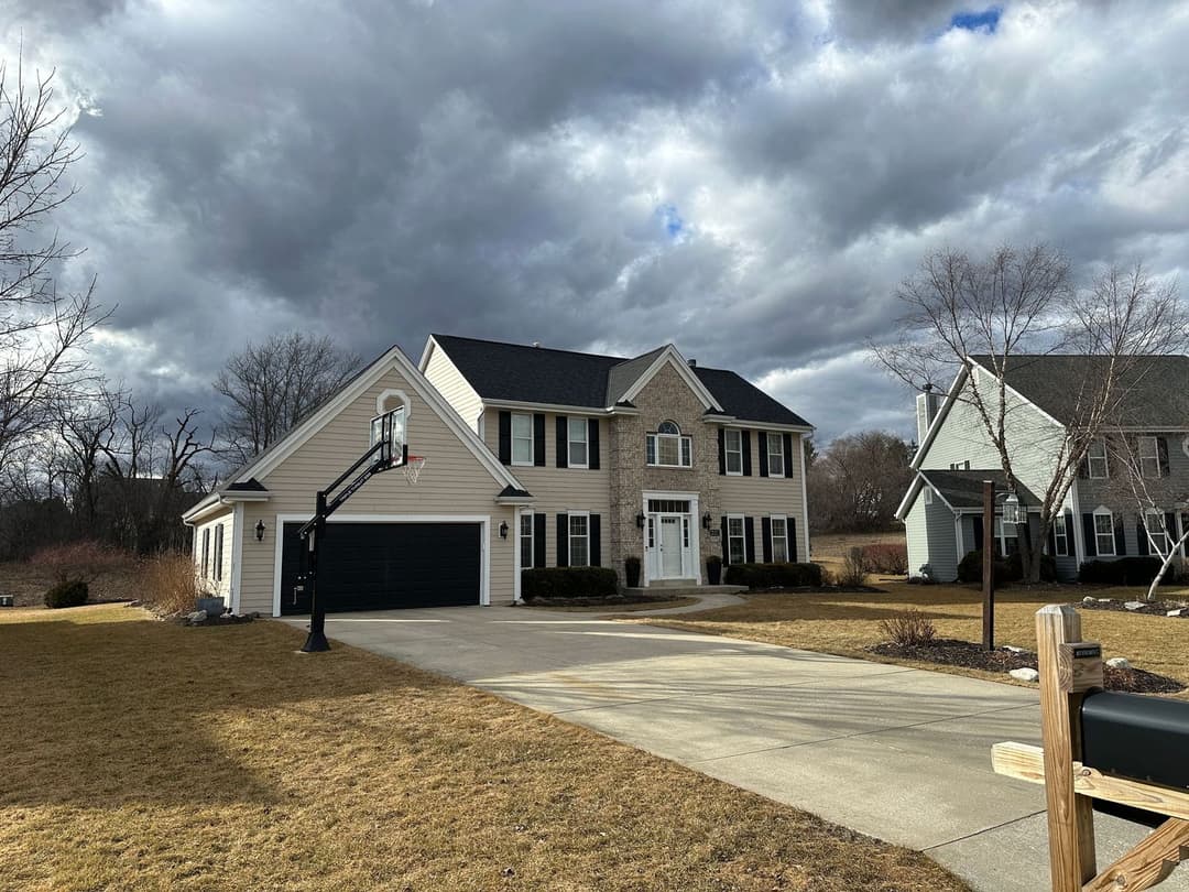 Two-story suburban home with basketball hoop, gray siding, and cloudy sky backdrop.