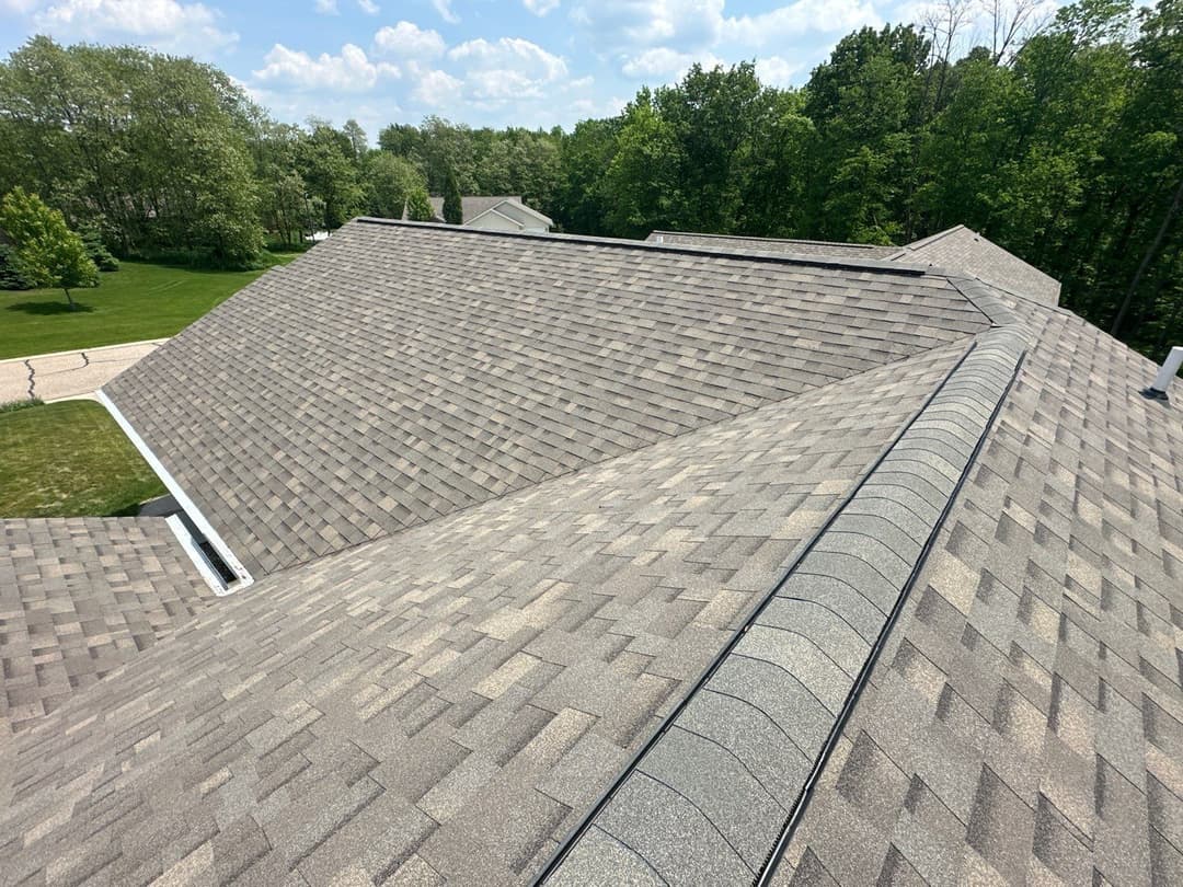 Aerial view of a shingled residential roof with forest and lawn in the background.
