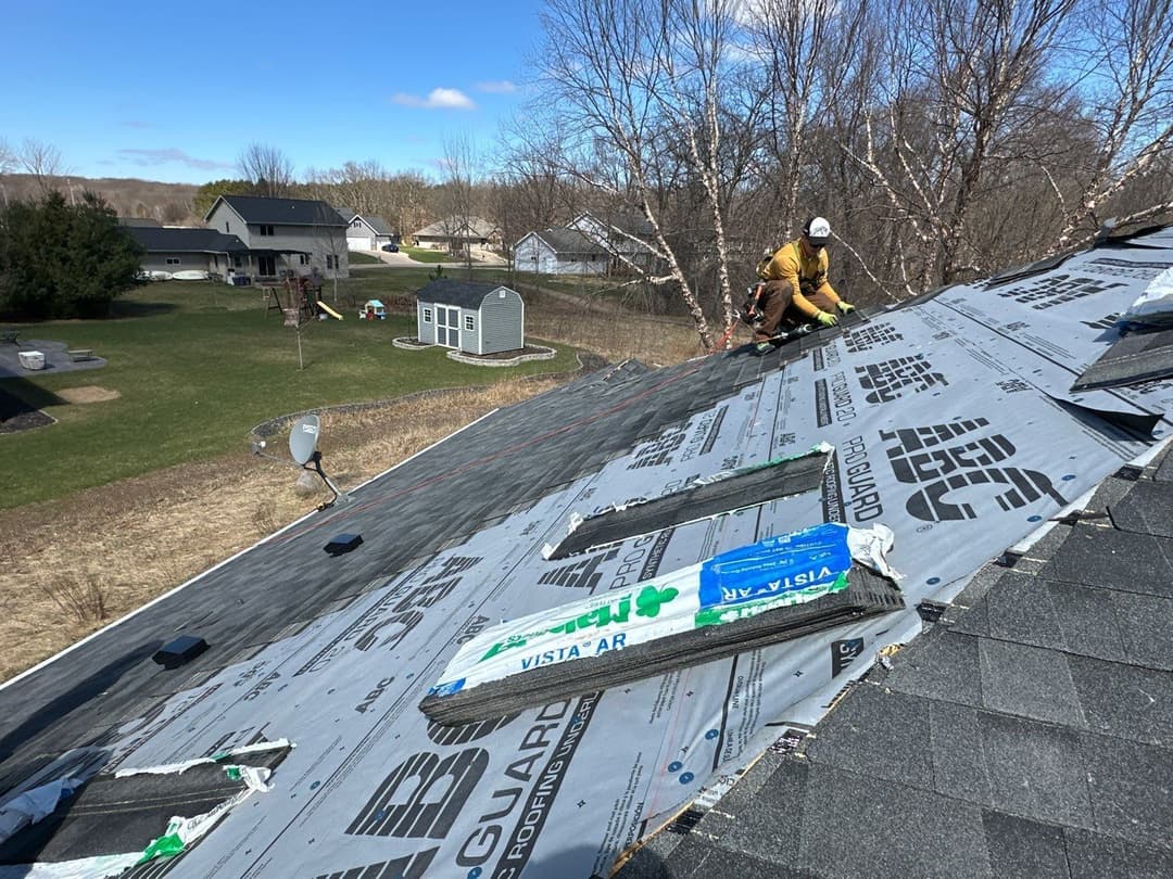 Roofing contractor installing shingles on a house, with surrounding residential area visible.