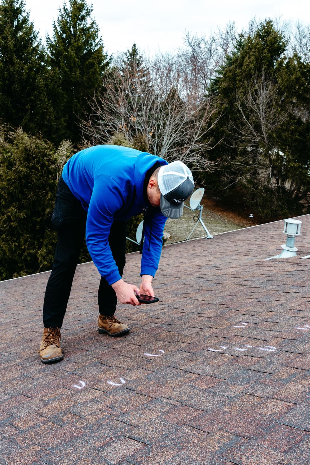 Man inspecting a roof with a smartphone, wearing a blue jacket and baseball cap.